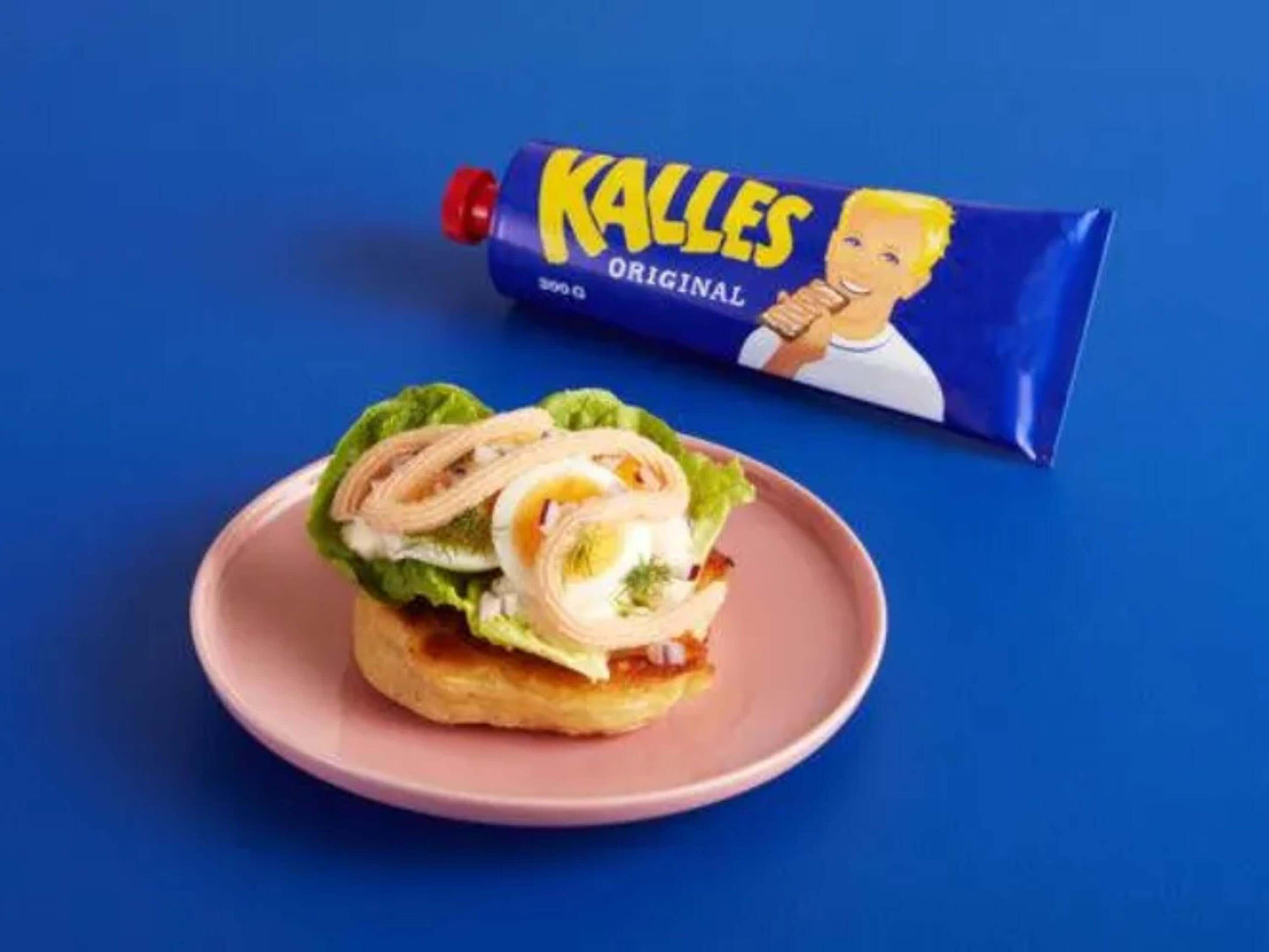 Kalles Kaviar Original tube beside a sandwich topped with lettuce and shrimp on a pink plate against a blue background.
